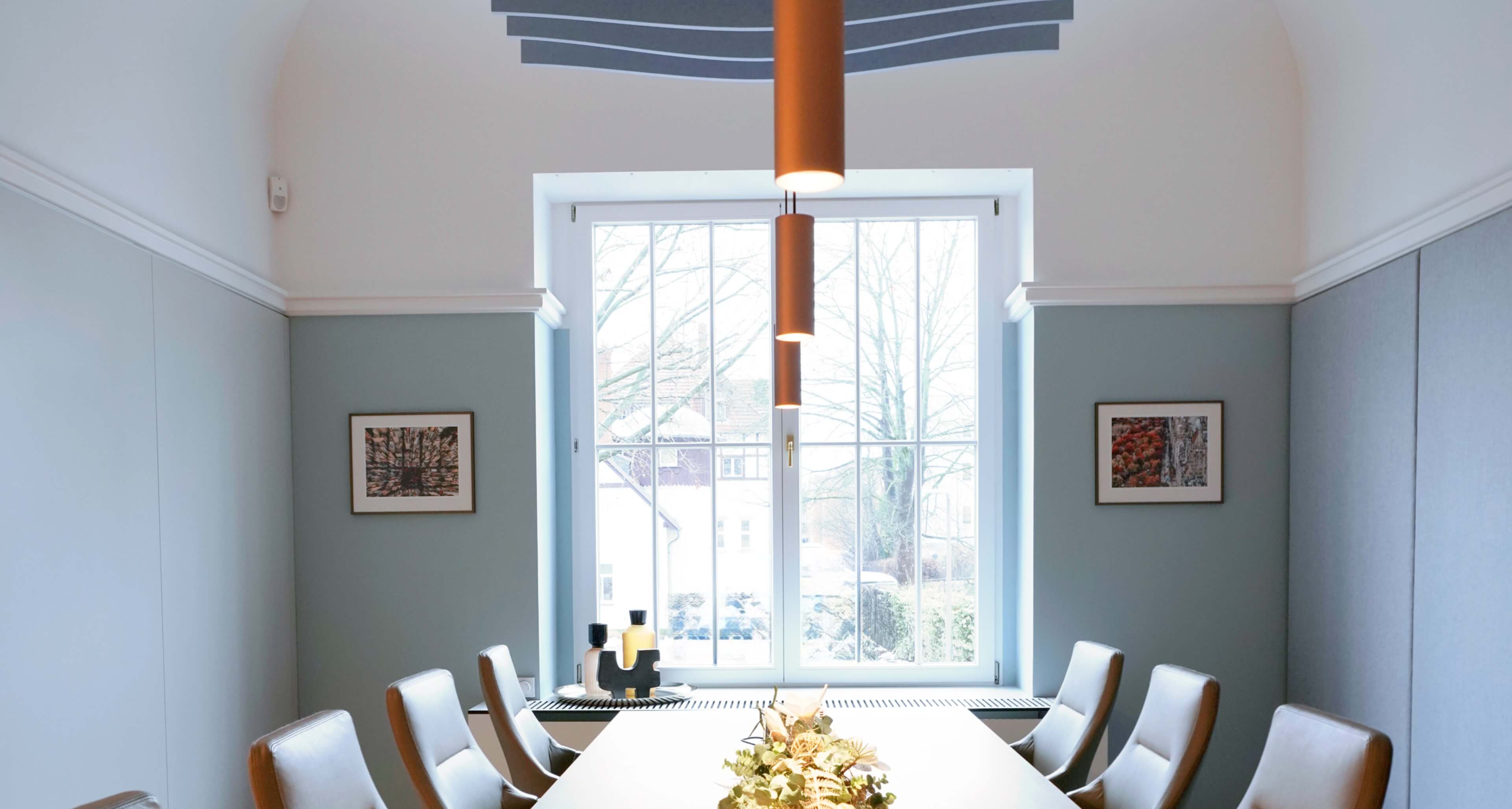 Heller, moderner Besprechungsraum mit langem Tisch, Stühlen, großem Fenster, orangefarbenen Pendelleuchten und gerahmter Kunst.