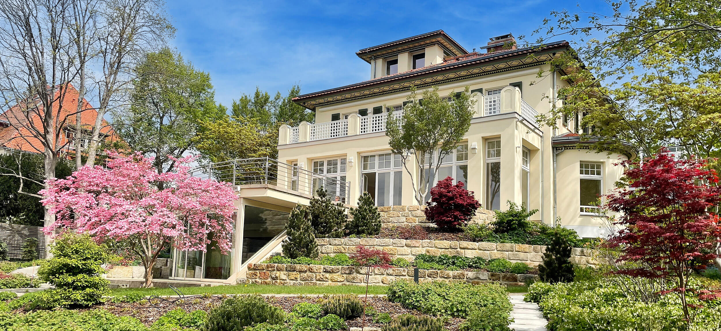Großzügige, mehrstöckige Stadtvilla mit heller Fassade, großen Fenstern und terrassiertem Garten mit blühenden Bäumen und Sträuchern an einem sonnigen Frühlingstag