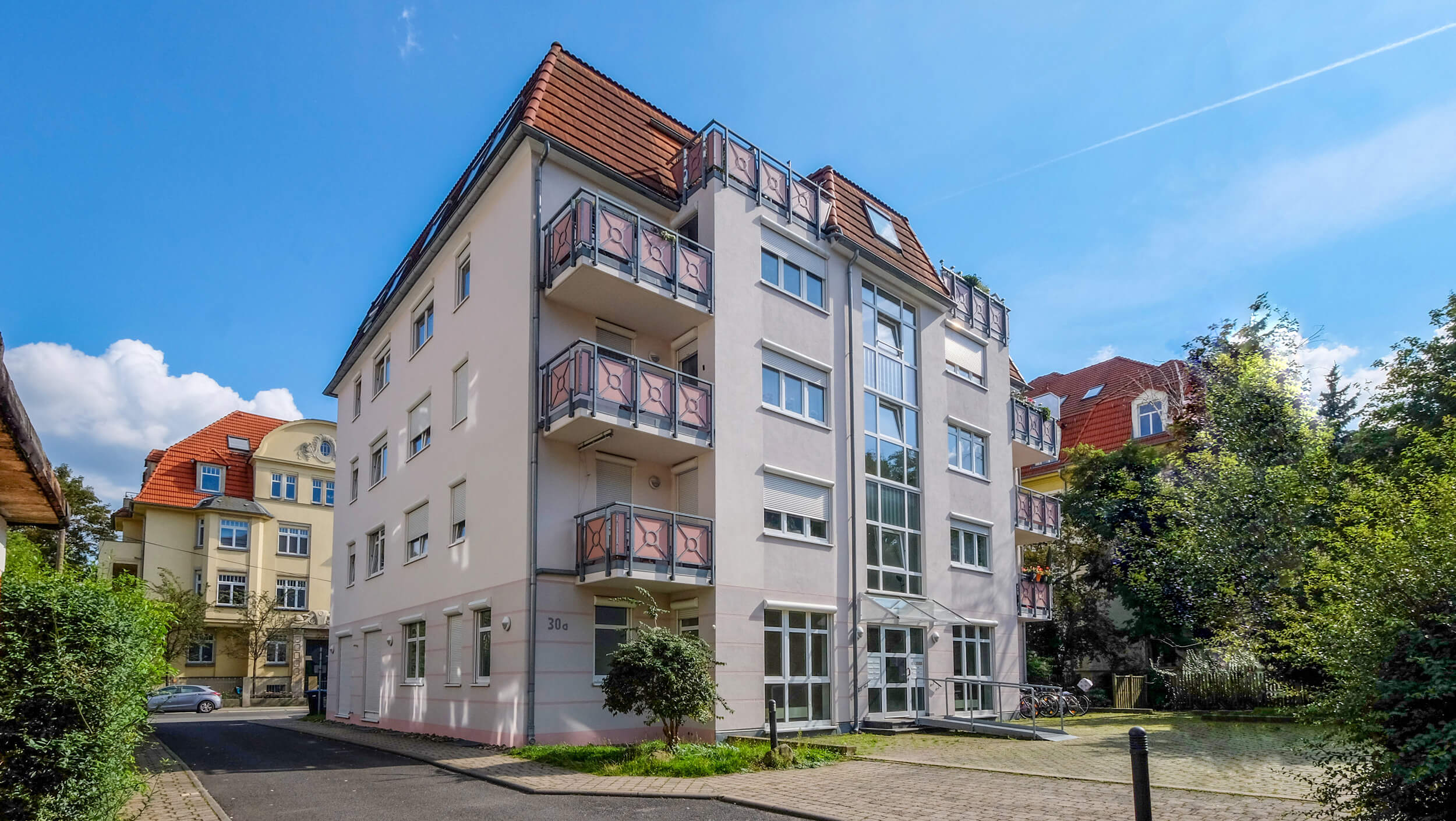 Modernes Mehrfamilienhaus mit heller Fassade, rotem Dach und mehreren Balkonen. Blauer Himmel, grüne Bäume.