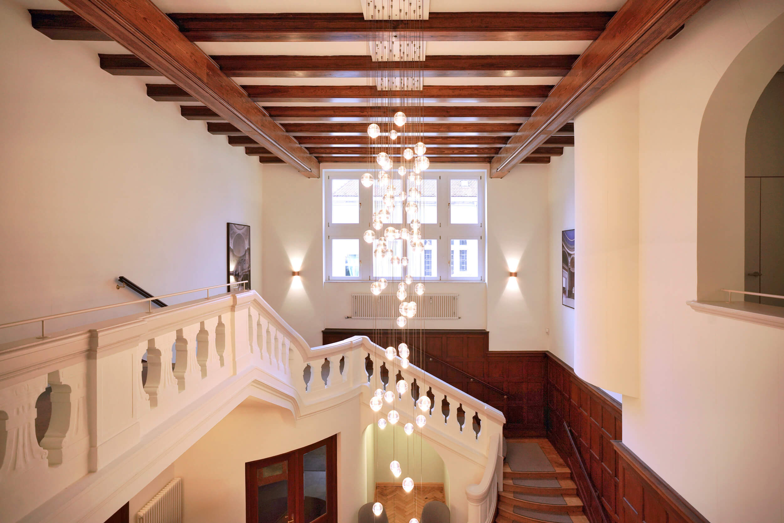Blick auf ein elegantes Treppenhaus mit weißer Balustrade, Holzbalken, großem Fenster und modernem Kugel-Kronleuchter.