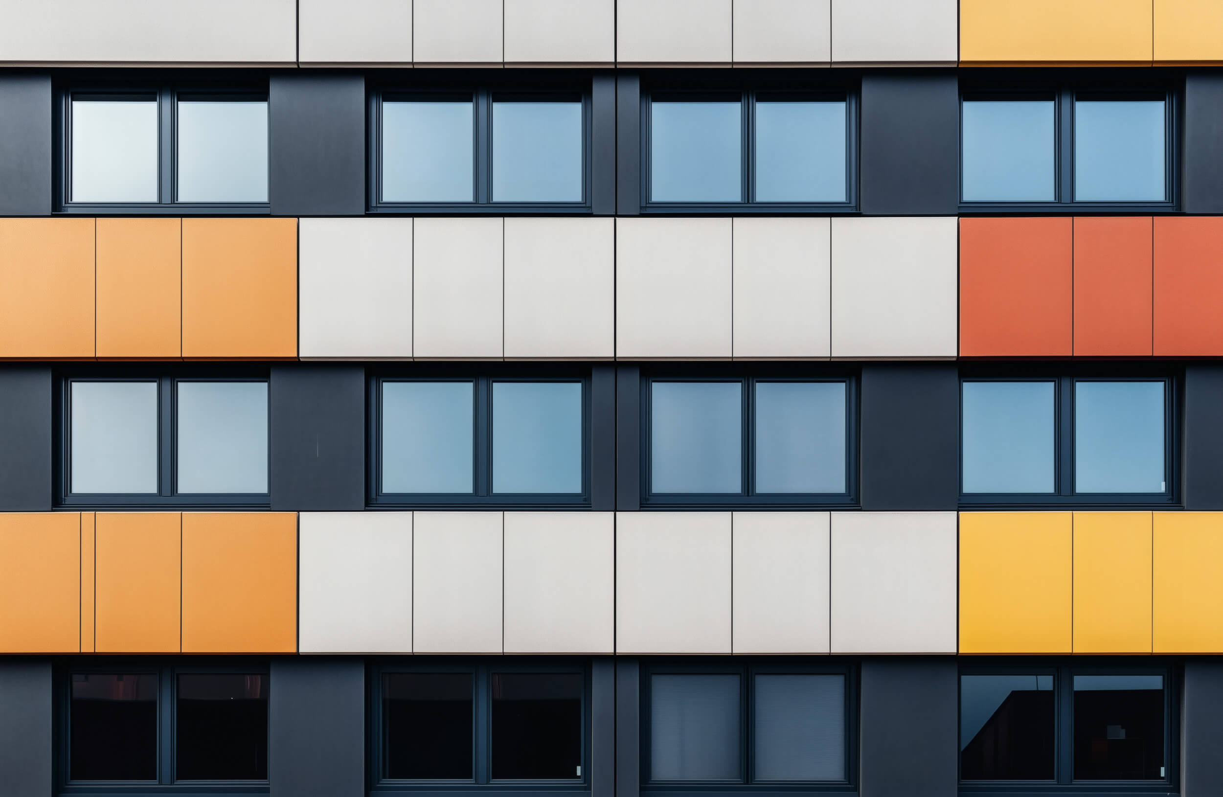 Geometrische moderne Gebäudefassade mit einem Muster aus Fenstern und farbigen Platten (Orange, Gelb, Grau, Weiß).