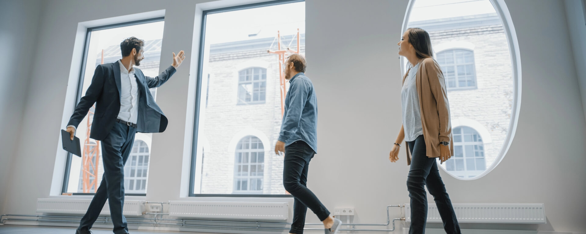 Drei Personen besichtigen eine helle, moderne Bürofläche mit großen Fenstern. Ein Mann im Anzug gestikuliert, während er spricht.
