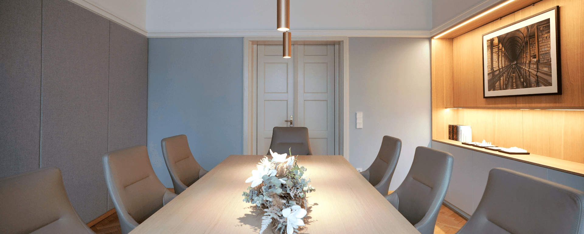 Eleganter Konferenzraum mit langem Holztisch, grauen Stühlen, modernen Leuchten, Akustikpaneelen und beleuchtetem Regal.