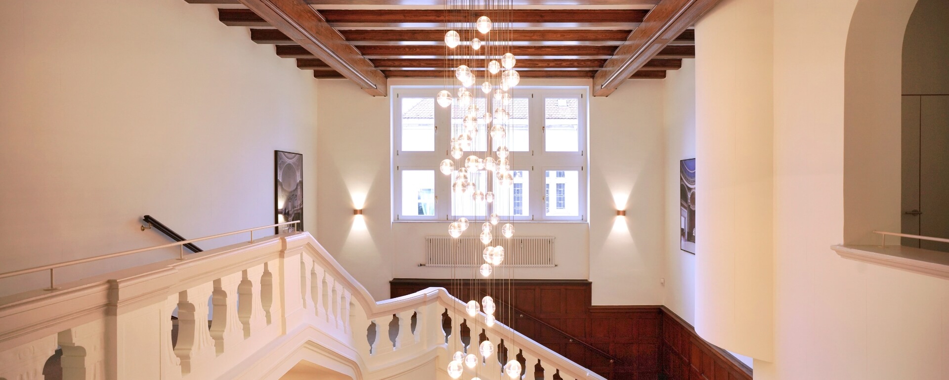 Blick auf ein elegantes Treppenhaus mit weißer Balustrade, Holzbalken, großem Fenster und modernem Kugel-Kronleuchter.