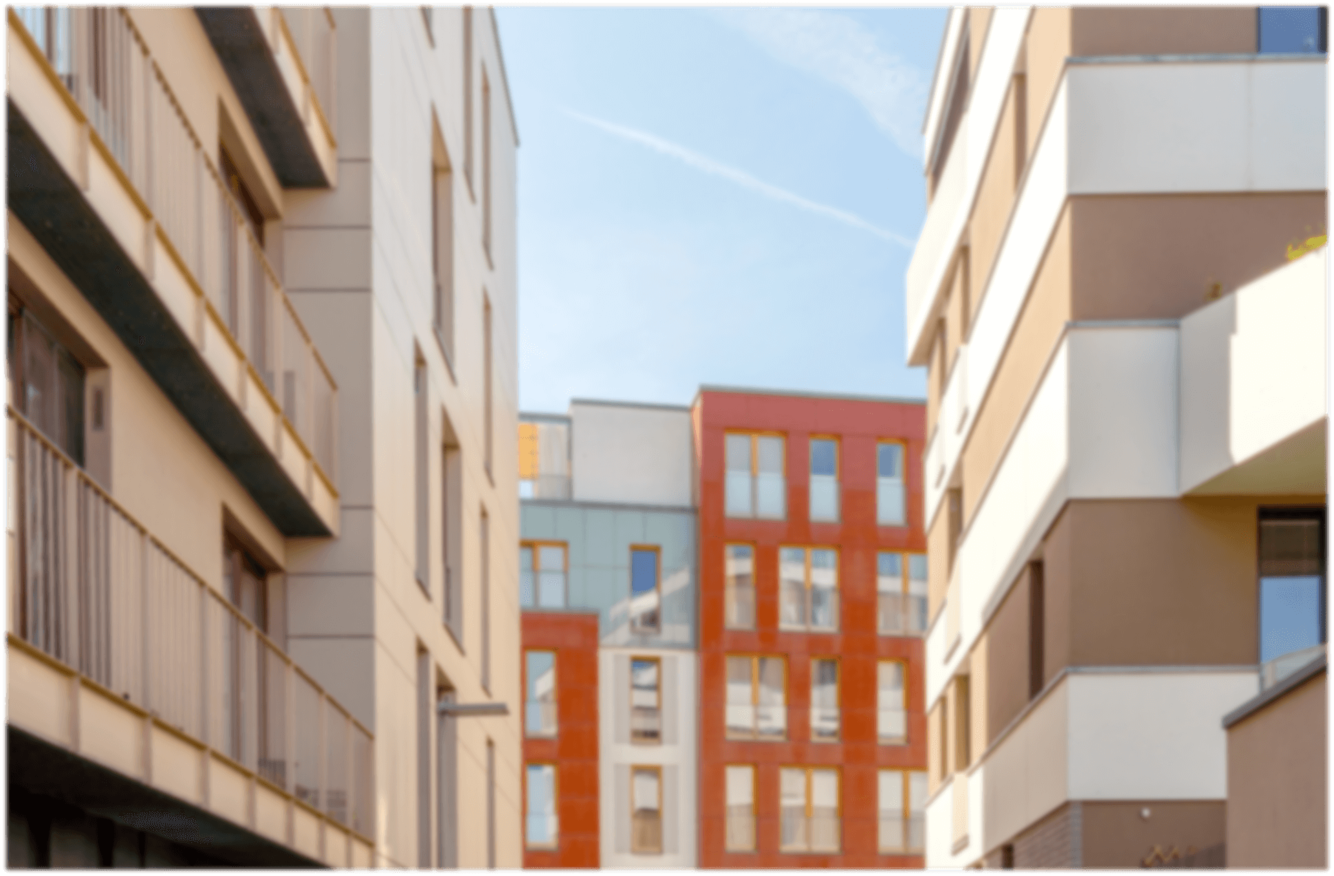 Verschwommene moderne Wohngebäude mit Balkonen in verschiedenen Farben, darunter rot-orange, unter hellem blauem Himmel.